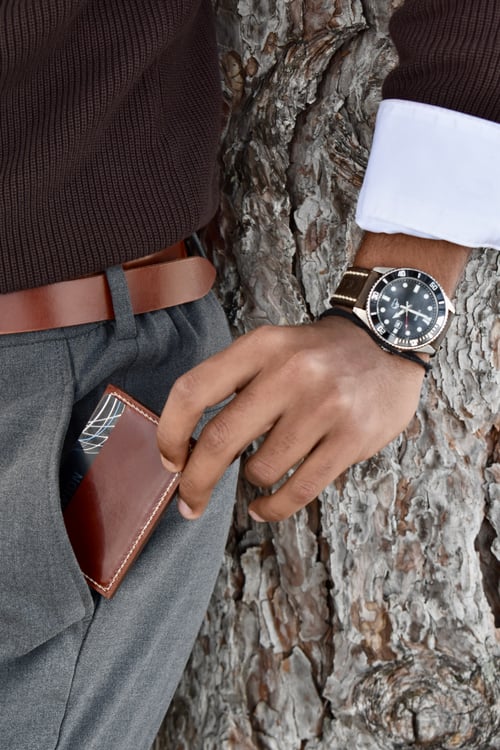 Close-up of a person standing against a tree trunk, wearing a brown sweater, gray trousers, and a brown belt, holding a brown leather card holder partially tucked into their pocket while wearing a wristwatch with a brown leather strap.