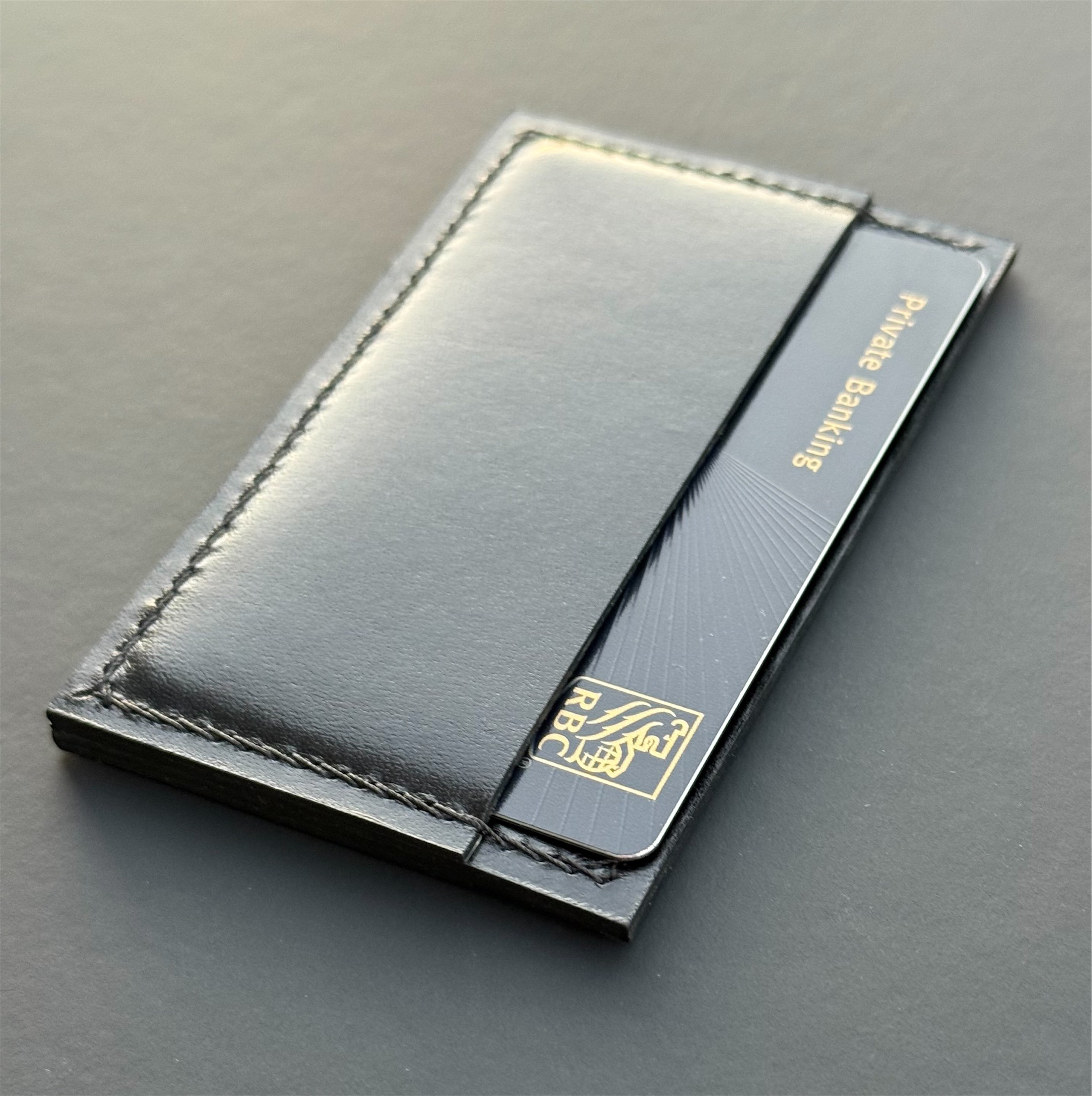 Black RBC Private Banking card partially inserted into a slim black leather card holder with stitched edges, photographed on a dark matte surface.